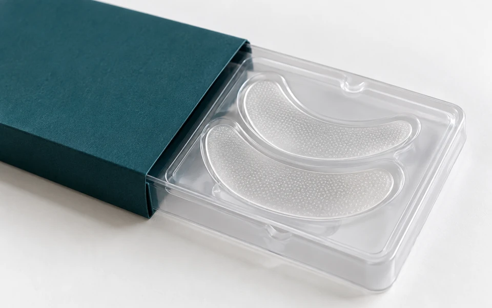 AI-generated illustrative product concept Paired crescent eye-patch prototypes in a clear sample blister and teal sleeve